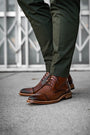 Brown leather shoes worn with green pants on a concrete surface.